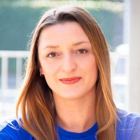 Edith Stecker lächelt in die Kamera. Sie hat braune Haare und ein blaues Oberteil und steht angelehnt an eine Wand.