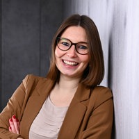 Maike Krebber Eine braunhaarige Frau mit Brille lehnt an einer Wand und lächelt in die Kamera
