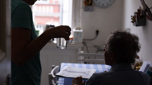 Eine Pflegerin reicht ihrem pflegebedürftigen Patienten bei einem Hausbesuch ein Glas Wasser.