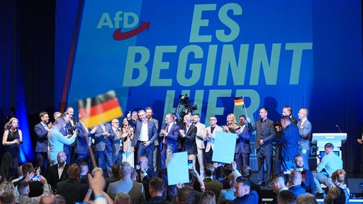 Auf einer Bühne stehen mehrere Menschen, einer schwenkt eine Deutschland-Flagge, im Hintergrund ist zu lesen "Es beginnt hier", links daneben das Loge der AfD