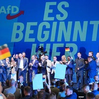 Auf einer Bühne stehen mehrere Menschen, einer schwenkt eine Deutschland-Flagge, im Hintergrund ist zu lesen "Es beginnt hier", links daneben das Loge der AfD