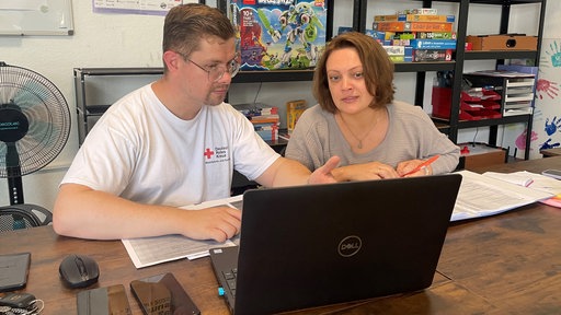 Dominik Meyer sitzt mit einer Klientin vor einem Laptop und erklärt ihr etwas. Vor ihnen liegen einige Zettel auf dem Tisch.Dom