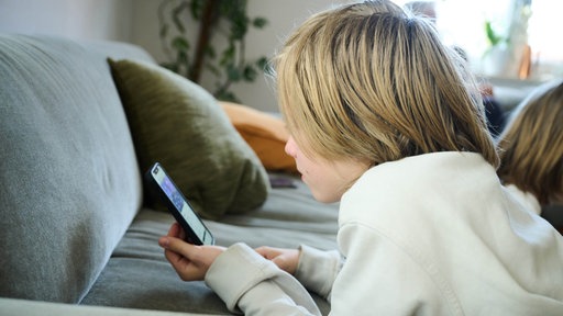 Ein Kind kauert vor einem Sofa und blickt auf ein Smartphone, man erkennt kein Gesicht