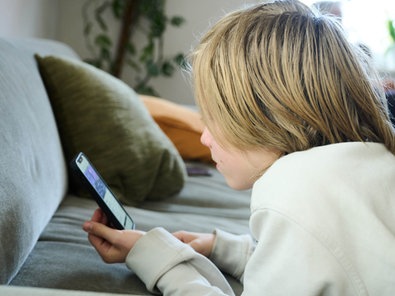 Ein Kind kauert vor einem Sofa und blickt auf ein Smartphone, man erkennt kein Gesicht