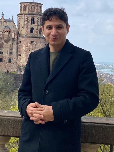 Migrantischer Mann steht mit Mantel vor einem Schloss und lächelt in die Kamera