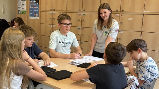 Zwei Medienscouts aus der zwölften Klasse der Gesamtschule Essen-Borbeck stehen an einem Tisch, an dem kinder aus der 5c sitzen