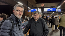 Zwei Männer stehen an einem bahnhof. Im Hintergrund laufen Menschen lang und man sieht eine Anzeigetafel.