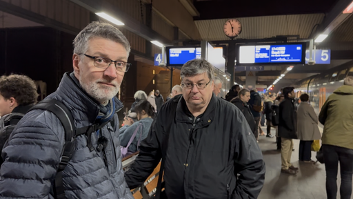 Zwei Männer stehen an einem bahnhof. Im Hintergrund laufen Menschen lang und man sieht eine Anzeigetafel.