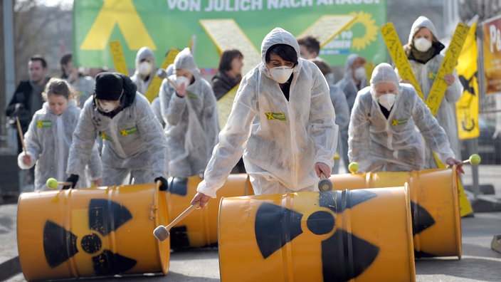 Aktivisten rollen Fässer mit Atomkraft-zeichen auf dem Boden, Sie tragen Schutzkleidung und Brillen. Im Hintergrund ein grünes Banner mit der Aufschrift" Kein Castor von Jülich nach Ahaus!"