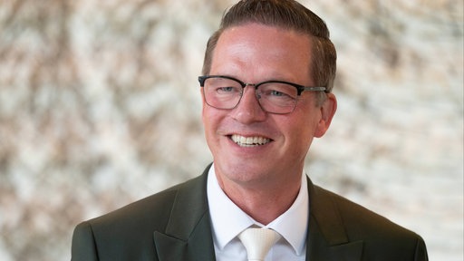 Gregor Golland - er trägt einen Anzug, hat kurze dunkelblonde Haare und trägt eine Brille. 