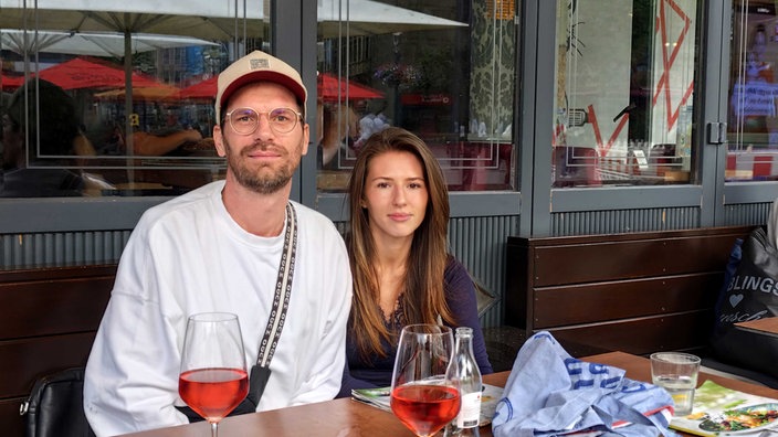 Ein Mann mit Brille und Kappe und eine dunkelhaarige junge Frau sitzen in einem Restaurant draußen.