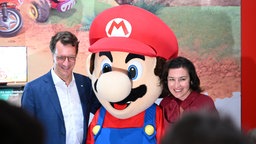 NRW-Ministerpräsident Hendrik Wüst und Bundesforschungsministerin Dorothee Bär stehen bei der Gamescom neben einer Werbepuppe für das Spiel Nintendo Mario Kart