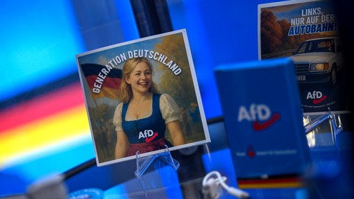 Sticker stehen auf einem Glastisch mit dem Slogan Generation deutschland. Auf dem einen ist eine blonde Frau in einem Dirndl zu sehen. Alle hhaben das AfD Logo drauf.