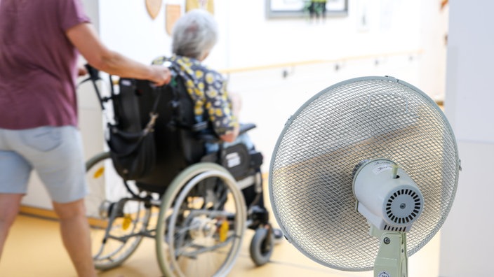 Eine Mitarbeiterin eines Seniorenheims schiebt eine Bewohnerin über den Flur, während ein Ventilator einen kühlen Luftzug erzeugt