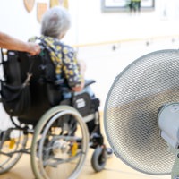 Eine Mitarbeiterin eines Seniorenheims schiebt eine Bewohnerin über den Flur, während ein Ventilator einen kühlen Luftzug erzeugt
