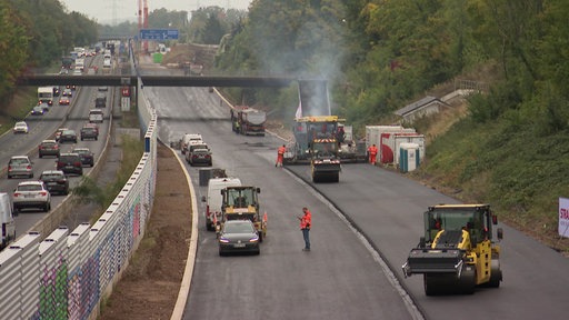 Baustelle an der A555, Bauarbeiter in orangener Warnkleidung und dampfende Baumaschinen, die neu asphaltieren.
