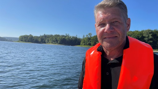 Fischereimeister Markus Kühlmann vom Ruhrverband mit orangener Schwimmweste an und im Hintergrund ein See.