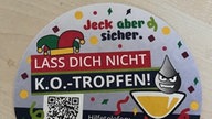Ein bunt bedruckter Bierdeckel liegt auf einem Tisch, auf ihm steht "Jeck aber sicher", dazu Infos zum Verhalten für Opfer von K.O.-Tropfen