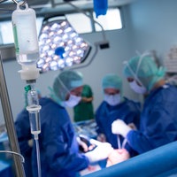Ärzte operieren in einem OP-Saal ein Knie, im Vordergrund sieht man den Tropf