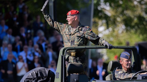 Brigadegeneral Hans-Dieter Müller steht in einem offenen Jeep mit camouflage Muster und winkt, im Hintergrund eine Menschenmenge.