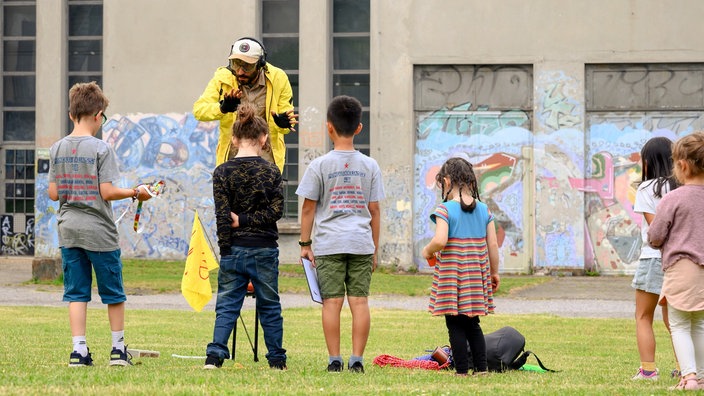 Ein Mann steht auf einer Wiese. Er trägt eine gelbe Windjacke, eine Schutzbrille und Ohrenschutz. Vor ihm stehen sechs Kinder. Er erklärt ihnen etwas.