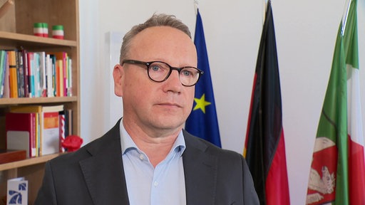 Benjamin Limbach im Interview, im Hintergrund ein Bücherregal und drei Fahnen: Europa-, Deutschland- und NRW-Fahne