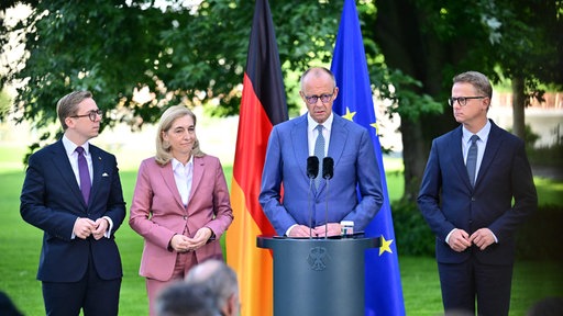 Friedrich Merz steht an einem Rednerpult, neben ihm Nina Warken, Caresten Linnemann und Philipp Amthor. Hinter ihnen die deutsche und die europäische Flagge.