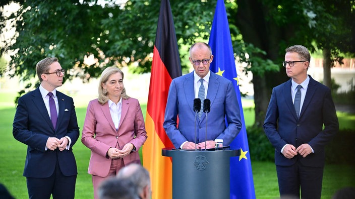 Friedrich Merz steht an einem Rednerpult, neben ihm Nina Warken, Caresten Linnemann und Philipp Amthor. Hinter ihnen die deutsche und die europäische Flagge.
