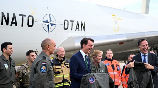 NRW- Ministerpräsident Hendrik Wüst und NATO-Generalsekretär Mark Rutte auf der Air Base in Geilenkirchen. Sie stehen vor einem Flugzeug von Leuten umgeben in Uniformen und halten Uniformen in der Hand. Beide lachen.