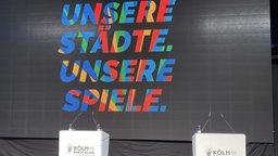 Auf einer Bühne stehen Rednerpulte, dahinter an einer schwarzen Wand steht bunt: "Unsere Städte. unsere Spiele."