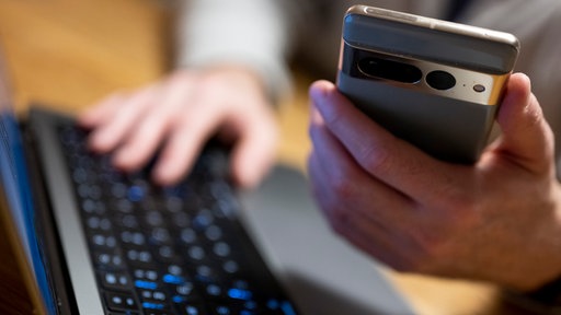 Im Vordergrund wird eine Handy in der Hand gehalten. Im Hintergrund tippt die selbe Person auf einer Tastatur.