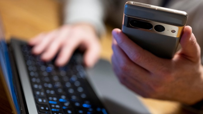 Im Vordergrund wird ein Handy in der Hand gehalten. Im Hintergrund tippt dieselbe Person auf einer Tastatur.