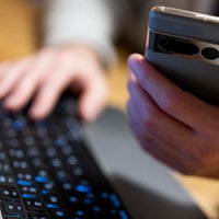 Im Vordergrund wird eine Handy in der Hand gehalten. Im Hintergrund tippt die selbe Person auf einer Tastatur.