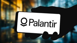 Name und Logo des Softwareunternehmens Palantir auf einem Smartphone, das jemand in der Hand hält. Dahinter bunte Lichter.