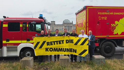 Sieben Männer halten ein schwarzgelbes Banner mit der Aufschrift "Rettet die Kommunen" hoch. Hinter ihnen stehen zwei Einsatzfahrzeuge der Feuerwehr und dahinter befindet sich das Reichstagsgebäude in Berlin.
