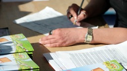 Frau sitzt an einem Tisch, in einer Hand einen Stift, und hält diesen über ein Blatt Papier
