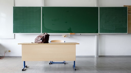 Schultafel in einer Klasse und davor steht ein Pult auf dem eine Tasche steht