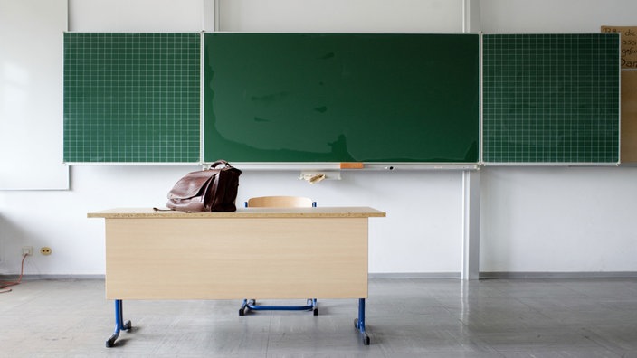 Schultafel in einer Klasse und davor steht ein Pult auf dem eine Tasche steht