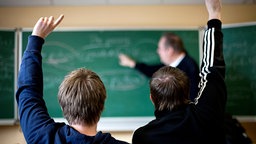 Zwei Schüler melden sich in einem Klassenraum der Schule, während der Lehrer an der Tafel steht (Foto vom 01.02.2011)