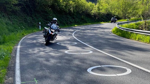 Blick auf die L218 im Kreis Düren, auf der Fahrbahn sind ellipsenförmige Markierungen, zwei Motorräder fahren in eine Kurve
