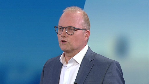 EIn Mann mit schütterem Haar, runder dunkler und Anzug vor einem blauen Hintergrund