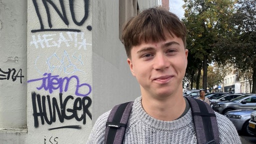 Ein junger Mann mit grauem Pulli und Rucksack schaut in die Kamera.