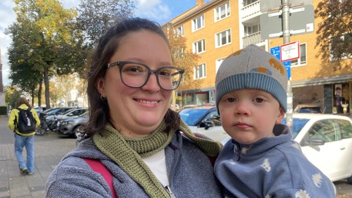 Eine Frau mit einer grauen Jacke und Brille schaut in die Kamera. Sie hat ein Kind mit blau-grauer Mütze auf dem Arm.