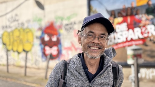 Ein Mann mit dunkler Cap, grauer Jacke und Brille schaut lachend in die Kamera. Im Hintergrund ist Graffiti unscharf an einer Wand.