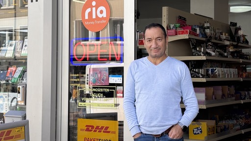 Ein Mann mit hellem Oberteil lehnt an der Tür eines Ladens und schaut in die Kamera. Im Hintergrund stehen Produkte im Laden und links neben ihm hängt ein rotes "Open"-Schild.