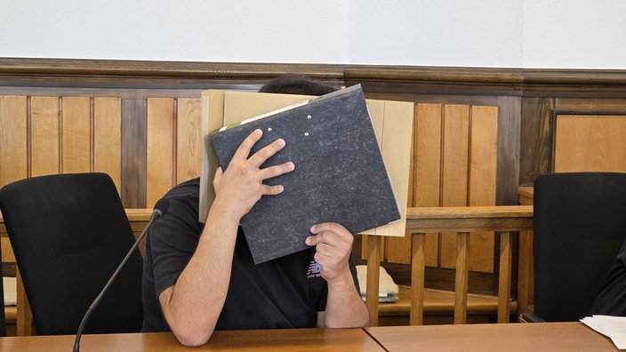 Vermeintlicher Notfallsanitäter sitzt im Gerichtssaal und hält sich einen Ordner vor sein Gesicht. 