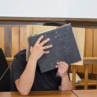 Vermeintlicher Notfallsanitäter sitzt im Gerichtssaal und hält sich einen Ordner vor sein Gesicht. 