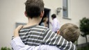 Zwei Kinder von hinten, die sich im Arm halten. Im Hintergrund sieht man die streitenden Eltern