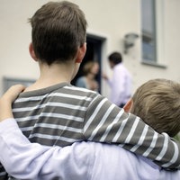 Zwei Kinder von hinten, die sich im Arm halten. Im Hintergrund sieht man die streitenden Eltern
