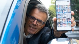 Uwe Baltner,  King of Carpool Karaoke, zeigt sein Handy aus derm Autofenster heraus und lacht. 
